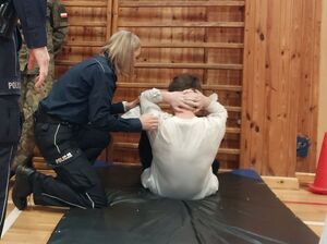 Zdjęcie w kolorze przedstawia ucznia szkół, wykonującego skłony z leżenia na materacu, po lewej policjantka udziela instrukcji.