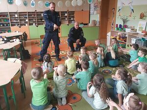 Zdjęcie w kolorze przedstawia przedszkolaków siedzących na kolorowym dywanie, na środku dwaj policjanci jeden stoi a drugi siedzi na małym krześle.