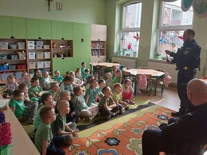 Zdjęcie w kolorze przedstawia przedszkolaków siedzących na kolorowym dywanie, na pierwszym planie dwaj policjanci jeden stoi a drugi siedzi na małym krześle.