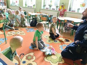 Zdjęcie w kolorze przedstawia przedszkolaków siedzących na kolorowym dywanie, na pierwszym planie na  środku troje przedszkolaków ćwiczy pierwszą pomoc na lalkach leżących przed nimi na dywanie, policjant udziela instrukcji.