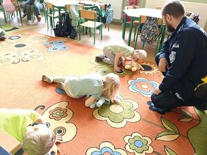 Zdjęcie w kolorze przedstawia policjanta,  który przykuca i instruuje dzieci przystawiające ucho do ust lalki leżącej na dywanie odwzorowując sprawdzanie oddechu.