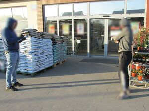 Zdjęcie w kolorze przedstawia dwóch mężczyzn przed wejściem do sklepu, mężczyzna po prawej stronie ma uniesione ręce wskazując dłońmi z nałożonymi kajdankami kierunek wejścia , drugi mężczyzna - policjant trzyma w dłoniach dokumenty i długopis.