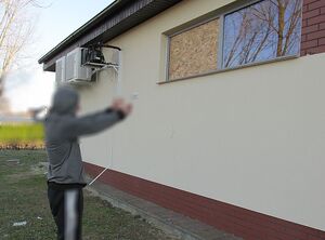 Zdjęcie w kolorze przedstawia mężczyznę, który wskazuje dłońmi, na których ma założone kajdanki na okno zasłonięte płytą ze sklejki.