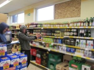 Zdjęcie w kolorze przedstawia dwóch mężczyzn we wnętrzu sklepu, mężczyzna po prawej stronie ma uniesione ręce wskazując dłońmi z nałożonymi kajdankami okno z wstawioną płyta ze sklejki pod którą są regały z napojami , drugi mężczyzna - policjant trzyma w dłoniach dokumenty i długopis.