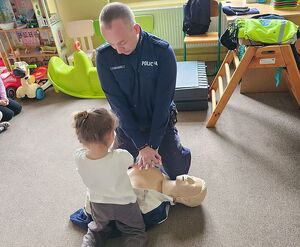 Zdjęcie w kolorze przedstawia policjanta klęczącego wspólnie z dziewczynką nad manekinem do nauki udzielania pierwszej pomocy.