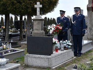 Zdjęcie w kolorze przedstawia dwóch policjantów w mundurach galowych stojących przed mogiłą, policjant po lewej trzyma kwiaty w kolorze białym i czerwonym