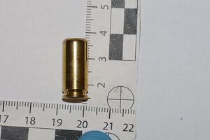 zdjecie w kolorze przedstawia mosiężna łuskę leżącą na białym tle przy skali pomiarowej