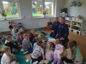 Zdjęcie w kolorze przedstawia grupę dzieci siedzących na podłodze razem z parą policjantów w sali przedszkolnej.