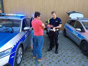 Zdjęcie w kolorze przedstawia policjanta stojącego pomiedzy radiowozem i policyjnym motocyklem, funkcjonariusz demonstruje odwiedzającym osobą dorosłym sprzęt.