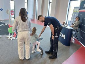 Zdjęcie w kolorze przedstawia policjanta trzymającego rowerek na który wsiada mała dziewczynka, obok stoi kobieta w tle miasteczko ruchu drogowego,