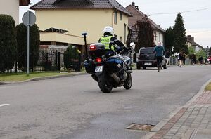 Zdjęcie w kolorze przedstawia policjanta kierującego motocyklem, który zabezpiecza peleton