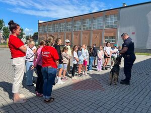 Zdjęcie w kolorze przedstawia grupę dzieci na placu zewnętrznym szkoły, po środku policjant z psem służbowym.
