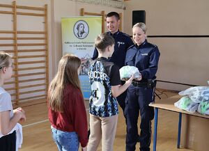 Zdjęcie przedstawia policjanta i policjantkę wręczających kaski dla dzieci w szkole.
