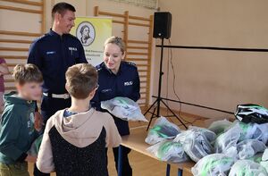 Zdjęcie przedstawia policjanta i policjantkę wręczających kaski dla dzieci w szkole.