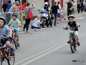 Uczestnicy wyścigu dziecięcego jadą do mety. W tle policjant ruchu drogowego na motocyklu zamyka skrzyżowanie