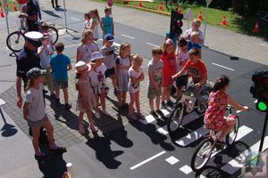 Dzieci na rowerach jeżdżą po miasteczku ruchu drogowego. Piesi czekają na zmianę światła przed przejściem dla pieszych.