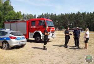 Policjant, Strażak i Komendant obozu stoją przy policyjnym radiowozie i wozie bojowym straży pożarnej.