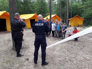 Policjant i Strażak stoją na polu namiotowym w lesie, przed nimi dzieci i młodzież stoją w szeregu. W tle żółte namioty.