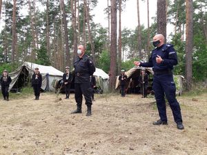 Policjant i Strażak stoją na polu namiotowym w lesie, wokół nich stoją harcerze.  W tle namioty i drzewa.