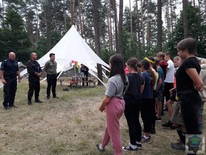 Policjant  w towarzystwie Strażaka i Komendanta Obozu Harcerskiego  stoją przed grupą dzieci . W tle drzewa i namioty.