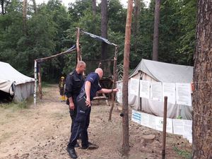 Policjant  i Strażak stoją przed regulaminami obozów, które wiszą na sznurkach między drzewami. Policjant wskazuje palcem na jedną z kart. Po obu stronach namioty, w tle drzewa.