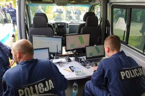 Centrum dowodzenia w jednym z radiowozów. Przed monitorami siedzi dwóch policjantów. Na monitorach widok map z zaznaczonymi sektorami do poszukiwań.