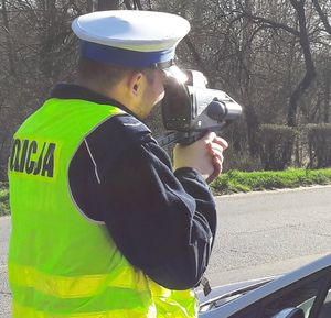 Policjant w mundurze i białej czapce mierzy prędkość za pomocą urządzenia pomiarowego.