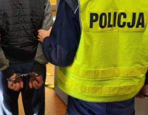 Mężczyzna w kamizelce odblaskowej z napisem policja trzyma innego mężczyznę pod ramię, który ma kajdanki na rękach.