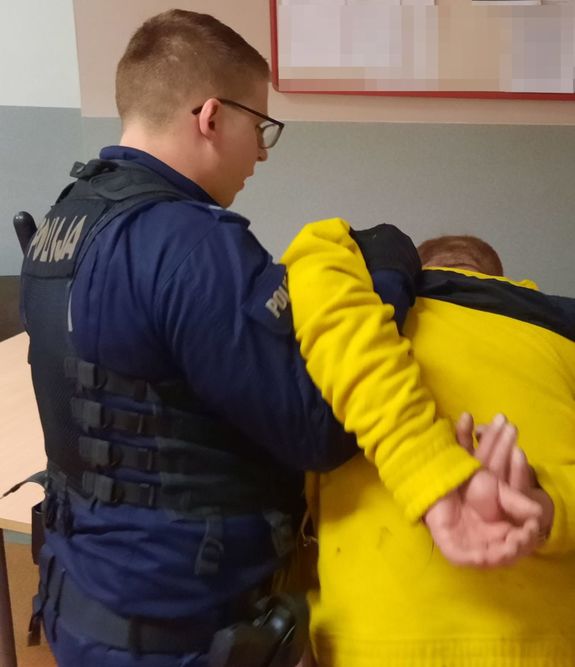 Policjant w mundurze przytrzymuje pod pachę mężczyznę w żółtej kurtce.