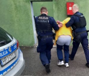 Dwóch umundurowanych policjantów prowadzi pod pachy mężczyznę w żółtej kurtce, obok stoi radiowóz.