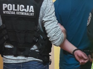 Mężczyzna w czarnej kamizelce z napisem policja trzyma pod rękę innego mężczyznę z kajdankami na rękach.