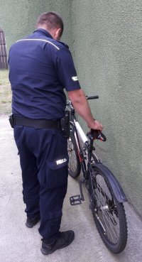 Mężczyzna ubrany w policyjny mundur stoi tyłem i trzyma rower za kierownicę.