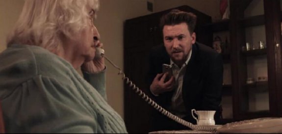 starsza kobieta trzyma słuchawkę telefonu stacjonarnego przy uchu, obok stoi mężczyzna z telefonem komórkowym w ręku
