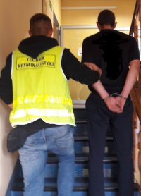 Mężczyzna w odblaskowej kamizelce z napisem technik kryminalistyki prowadzi po schodach drugiego mężczyznę, który ma założone kajdanki na ręce trzymane z tyłu.