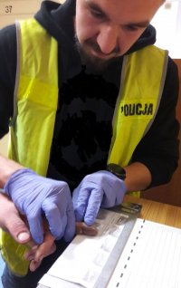 Mężczyzna w kamizelce odblaskowej z napisem policja pobiera odciski palców na białą kartkę od drugiego mężczyzny.