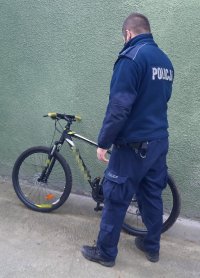 Policjant w mundurze stoi przy rowerze górskim na tle zielonej ściany.