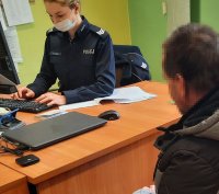 Policjantka w mundurze pisze na klawiaturze komputera, przed nią siedzi mężczyzna w szarej kurtce.