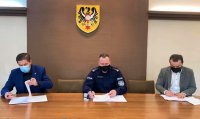 Trzech mężczyzn siedzi za dużym stołem i podpisują dokumenty, jeden z nich ma na sobie policyjny mundur.