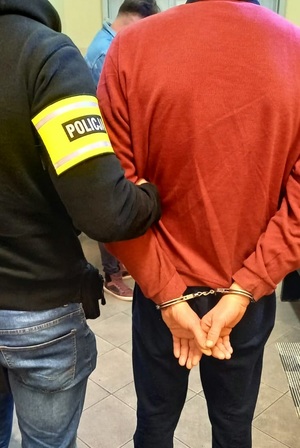 stojący tyłem mężczyzna z założonymi na ręce kajdankami, obok stojący policjant z opaska na ręce z napisem policja