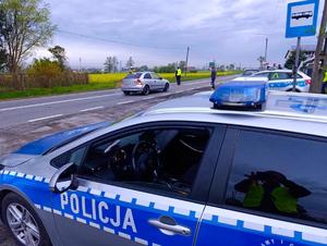 na pierwszym planie oznakowany radiowóz w dalszym planie policjanci umundurowani zatrzymujący na drodze inne pojazdy
