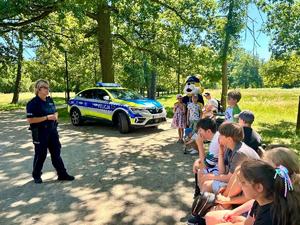 grupa dzieci słuchająca prelekcji policjantki w tle radiowóz