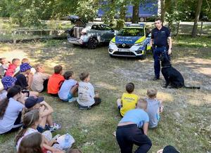 policjant z psem służbowym prowadzi prelekcję, na pierwszym planie siedzą dzieci