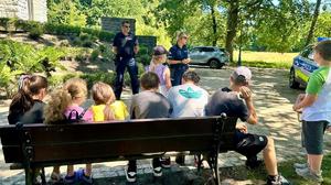 policjanci prowadzący prelekcje, w tle siedzące na ławce dzieci