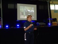 policjantka prowadząca spotkanie z hali sportowej, za nią wyświetlana prezentacja ze zdjęciami z wypadków drogowych