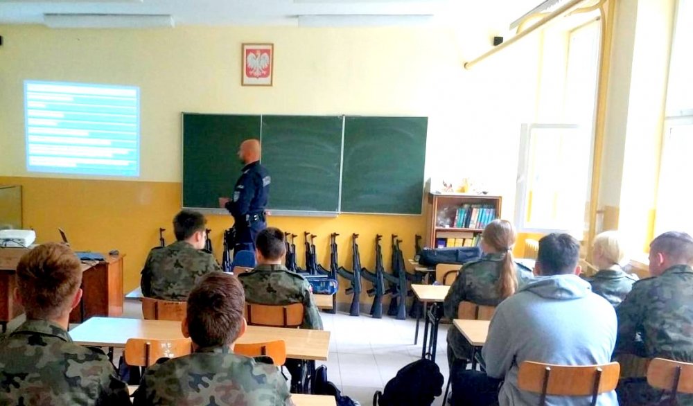 w klasie przy ławkach siedzą uczniowie naprzeciwko stoi policjant