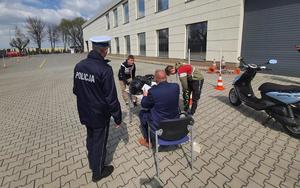 policjant stoi na placu obok ludzie siedzą na krzesłach