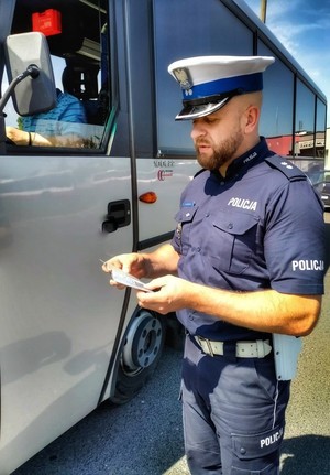 Zdjęcie przedstawia policjanta kontrolującego dokumenty, który stoi przy autokarze.