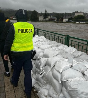 Zdjęcie przedstawia policjanta, idącego wzdłuż wału rzeki, obłożonego piaskiem.