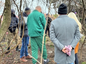 Zdjęcie przedstawia osoby stojące na łące, jedna z nich jest ubrana w zielony kombinezon i ma kajdanki na rękach i nogach.