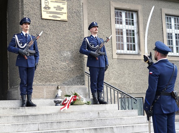 Policjant oddaje honor, w tle na baczność stoi dwóch policjantów.
